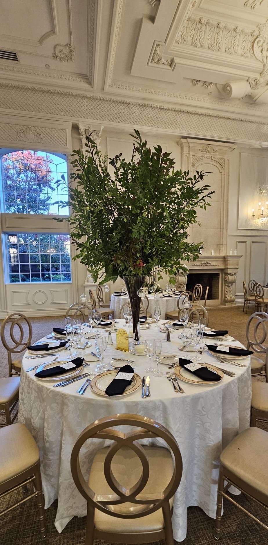 Tall branching greenery centerpiece above a black-and-ivory place setting in a light-filled reception room