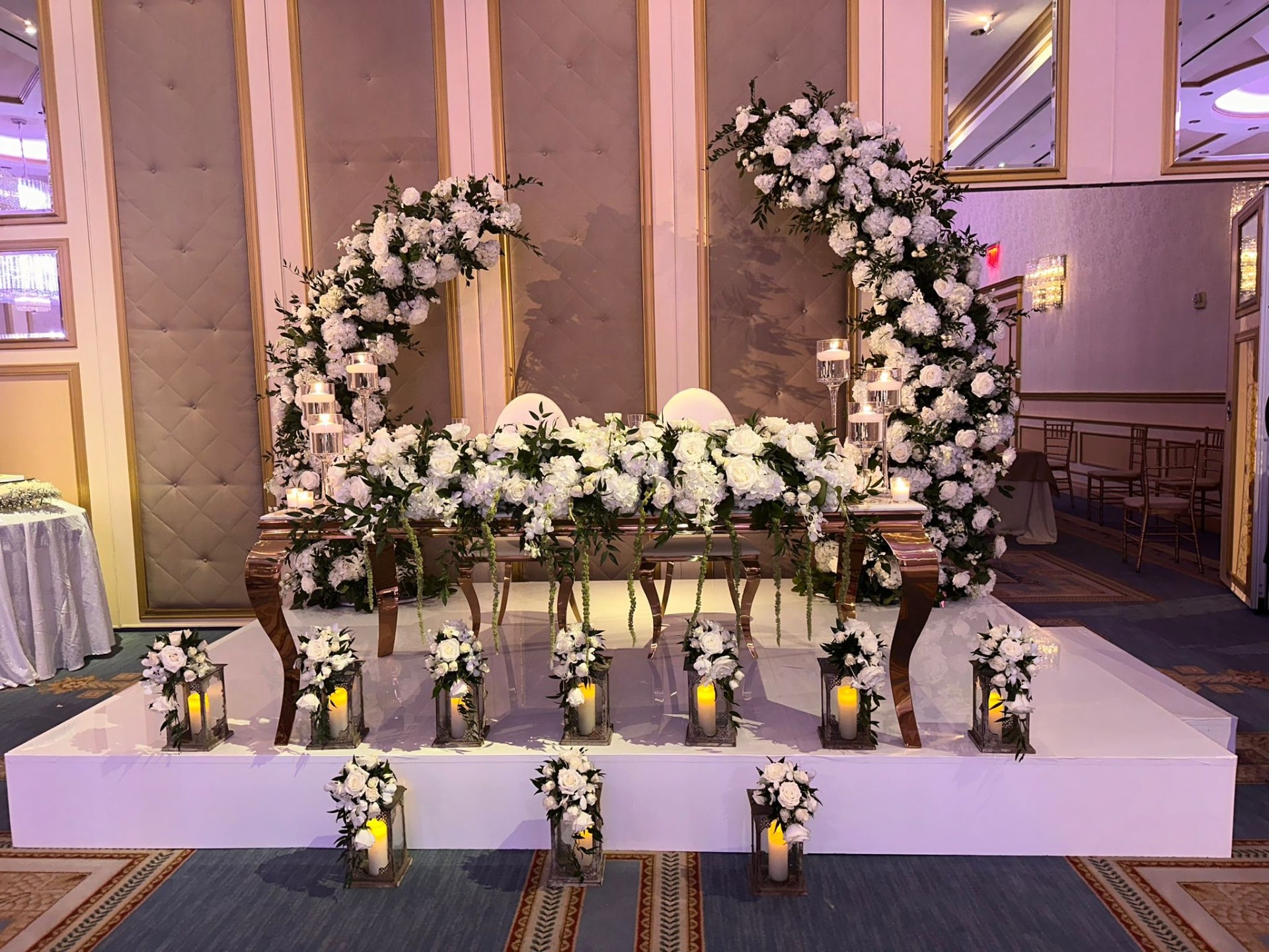 Sweetheart table beneath twin white rose and hydrangea arches, lined with lanterns, at a New Jersey ballroom wedding